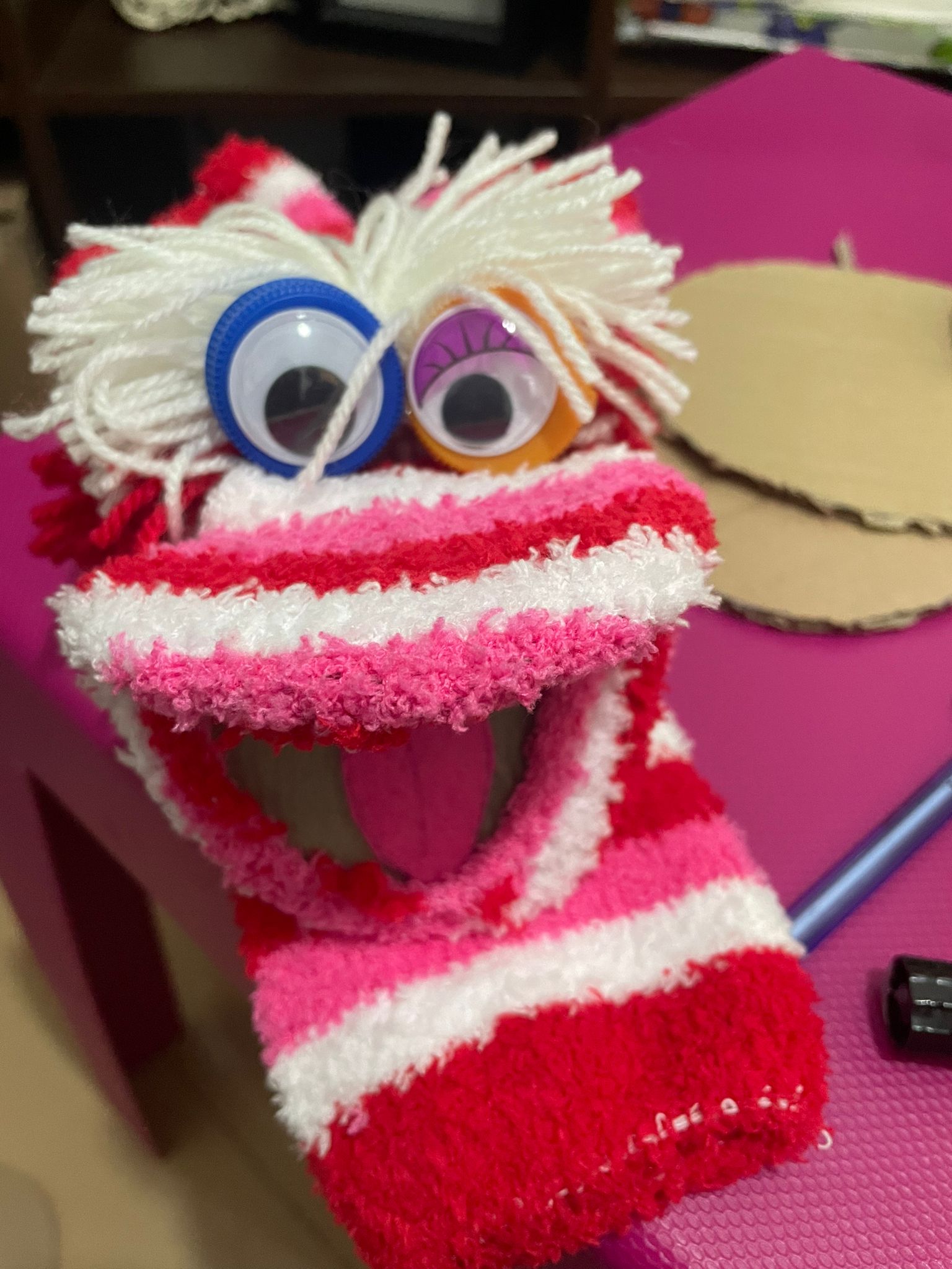 A imagem mostra uma marioneta de meia já pronta, com um ar simpático e divertido. Está construída com uma meia felpuda com listas largas de cor branca, rosa e vermelha. O cabelo é de cor branca e os olhos assimétricos, grandes, o esquerdo com contorno de cor azul e o direito com contorno inferior a laranja e a pálpera violeta. A marioneta tem a boca aberta e vê-se a língua rosa no interior.